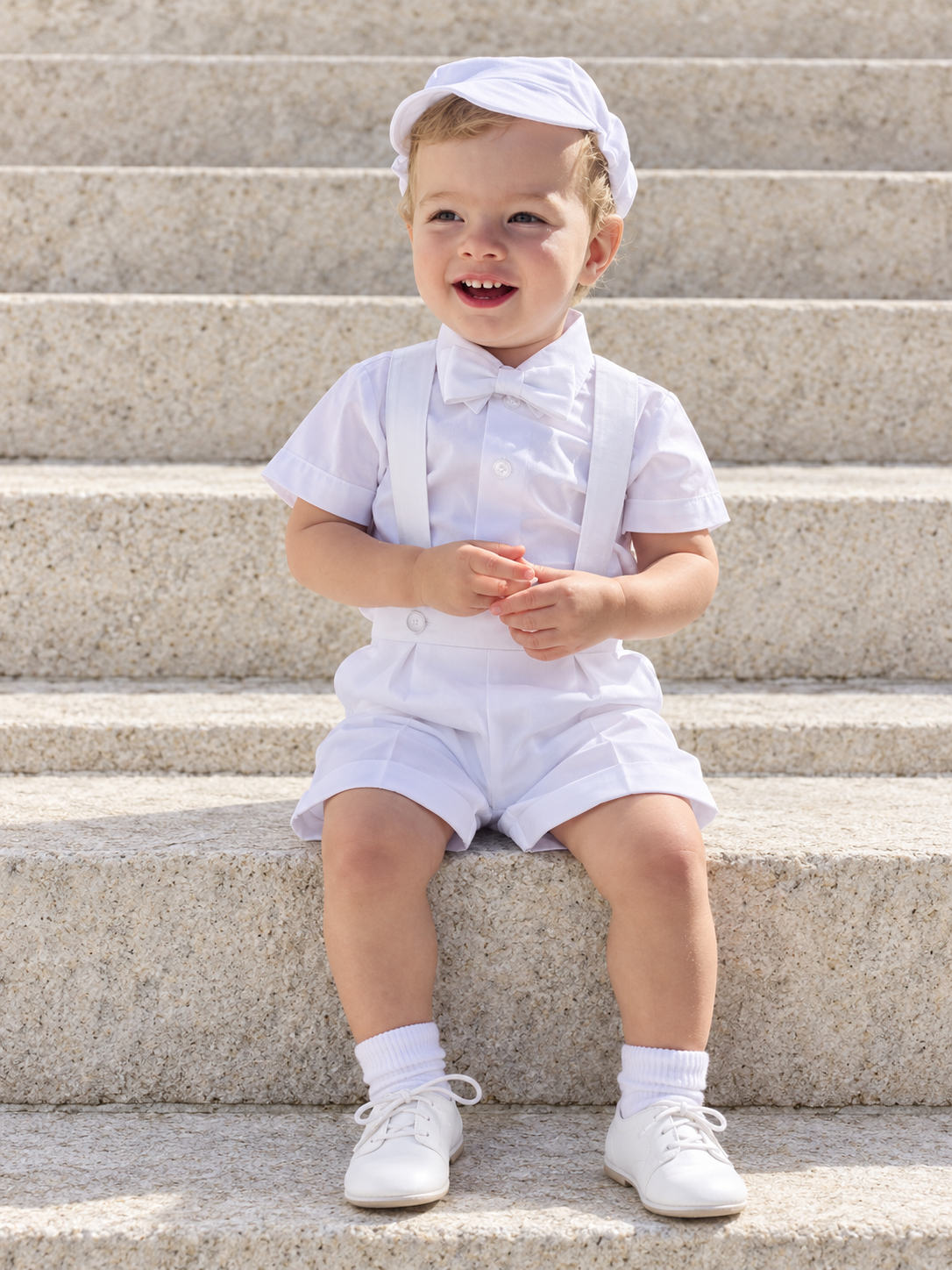 Toddler boy seated on stone steps wearing the white 850 baptism outfit with short-sleeve shirt, bow tie, suspender shorts and matching cap.