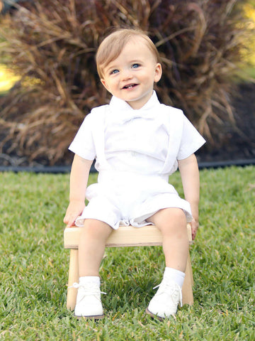 Baby boy seated outdoors wearing the white Blake baptism outfit with short-sleeve shirt, bow tie and suspender shorts.