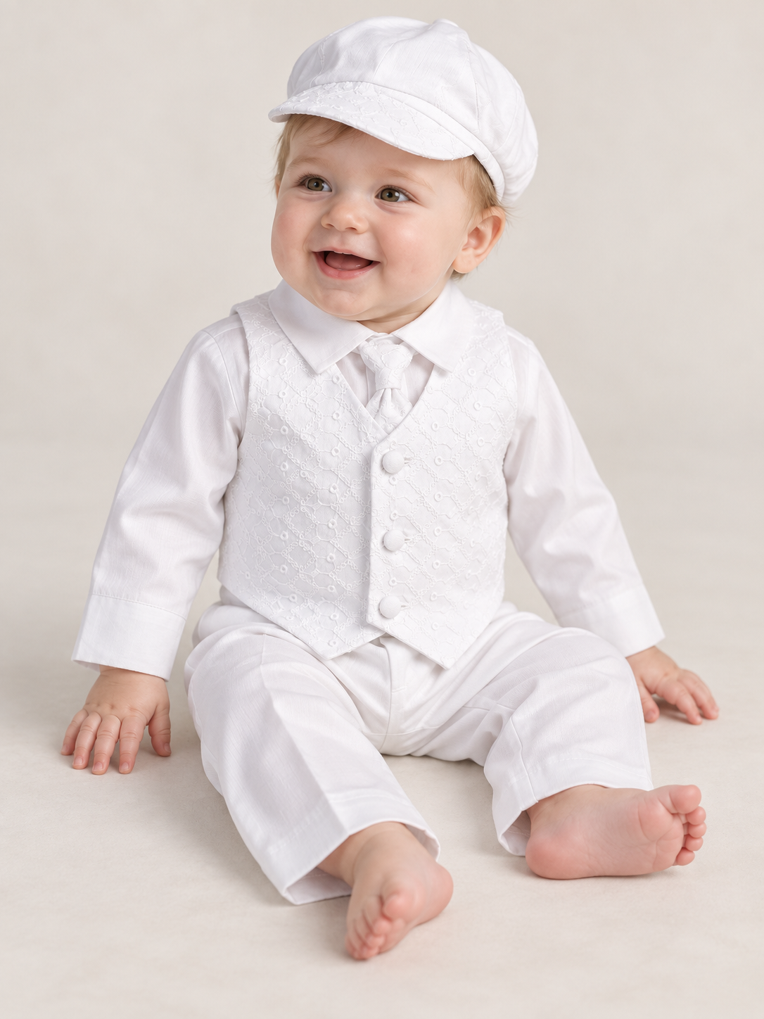 Seated baby boy wearing the white Corey baptism outfit with embroidered vest, matching necktie and cap, long-sleeve shirt and trousers.