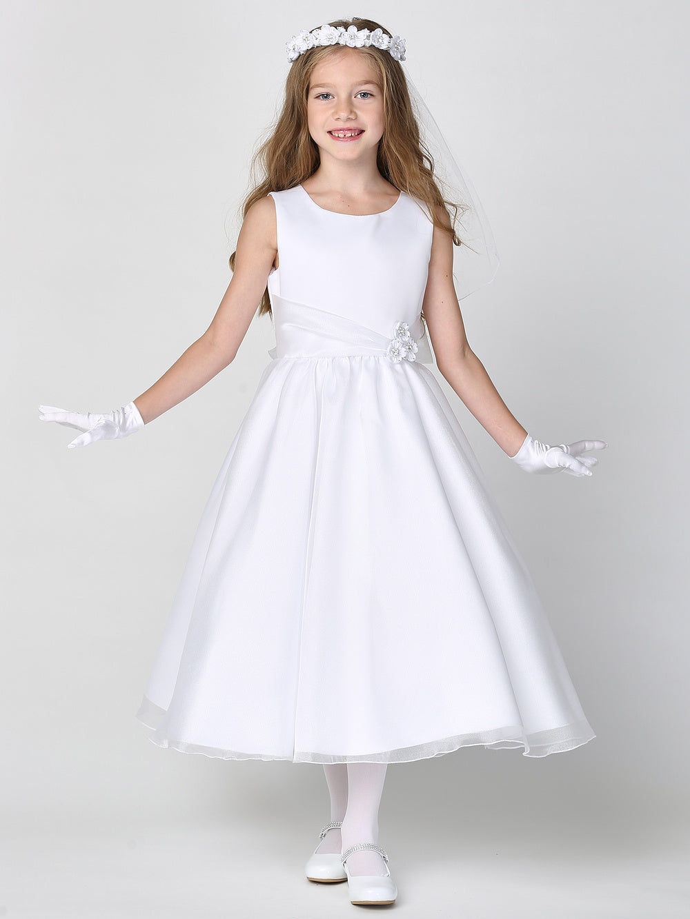 A girl wearing an elegant white First Communion Dress with a satin bodice and crystal organza skirt.