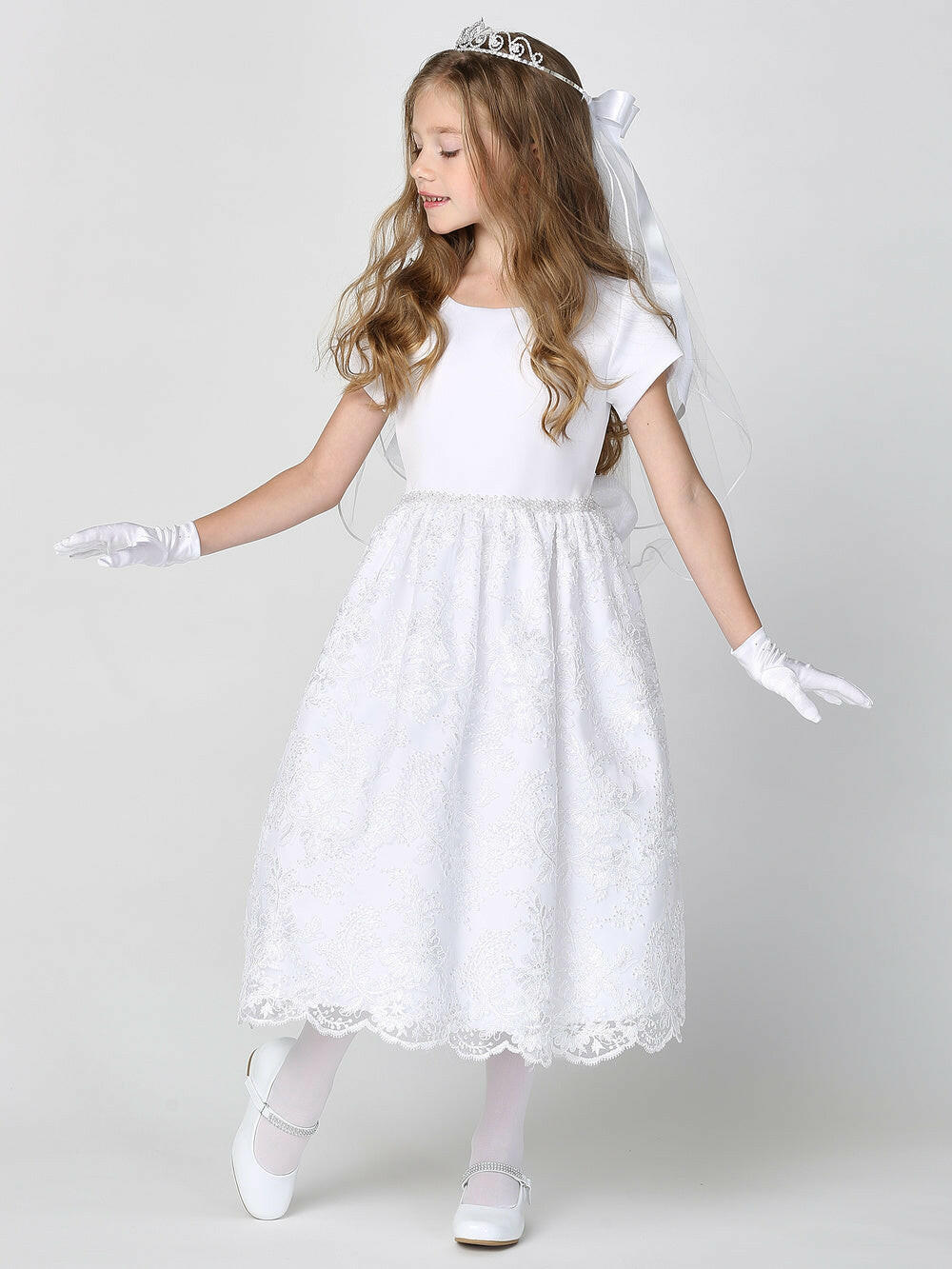 A girl wearing a beautiful white First Communion Dress with a satin bodice, corded lace skirt, and embroidered tulle with sequins. 