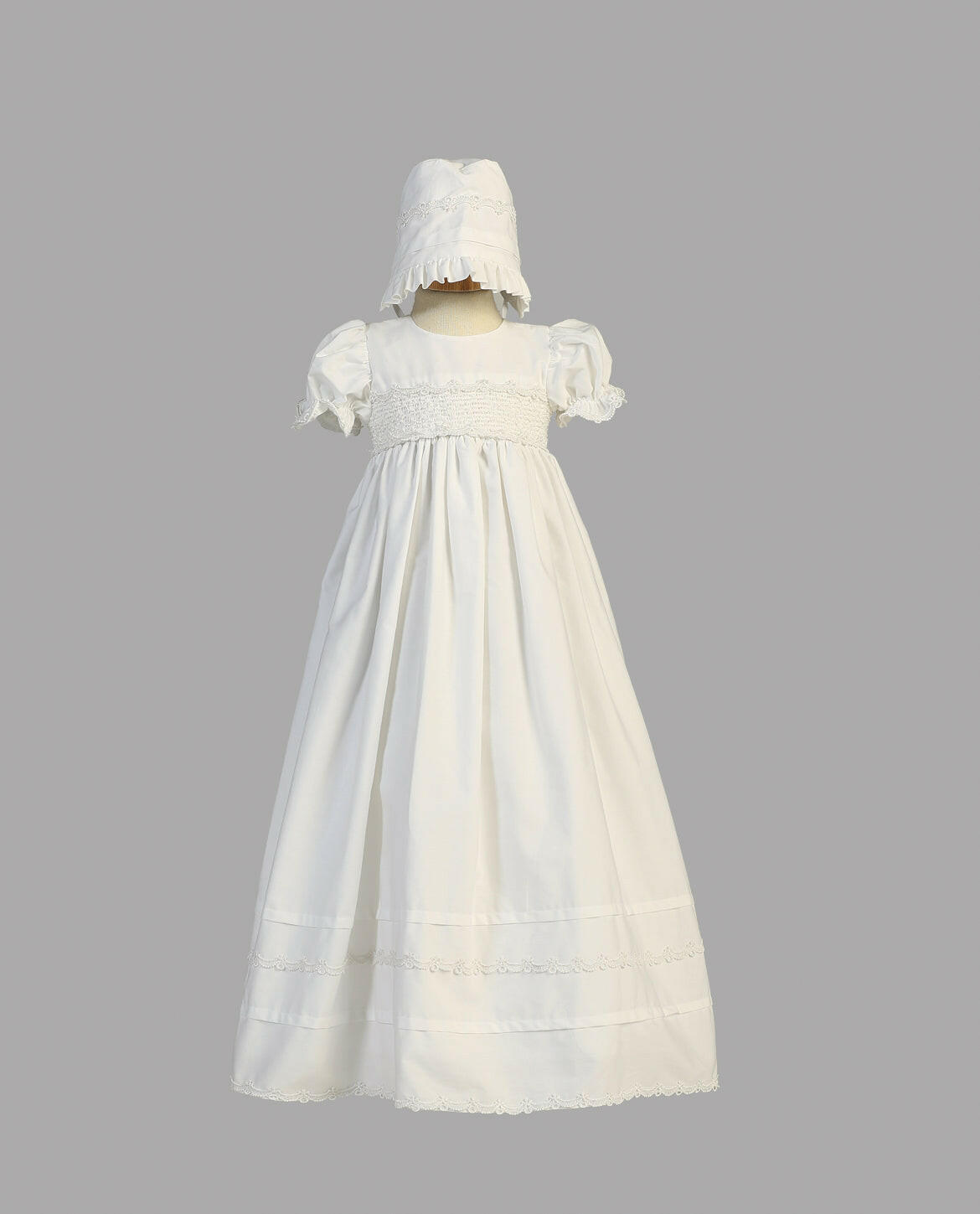 Girls White Baptism Gown Marie displayed on mannequin with bonnet, showcasing puff sleeves, lace detailing, and full-length cotton skirt.