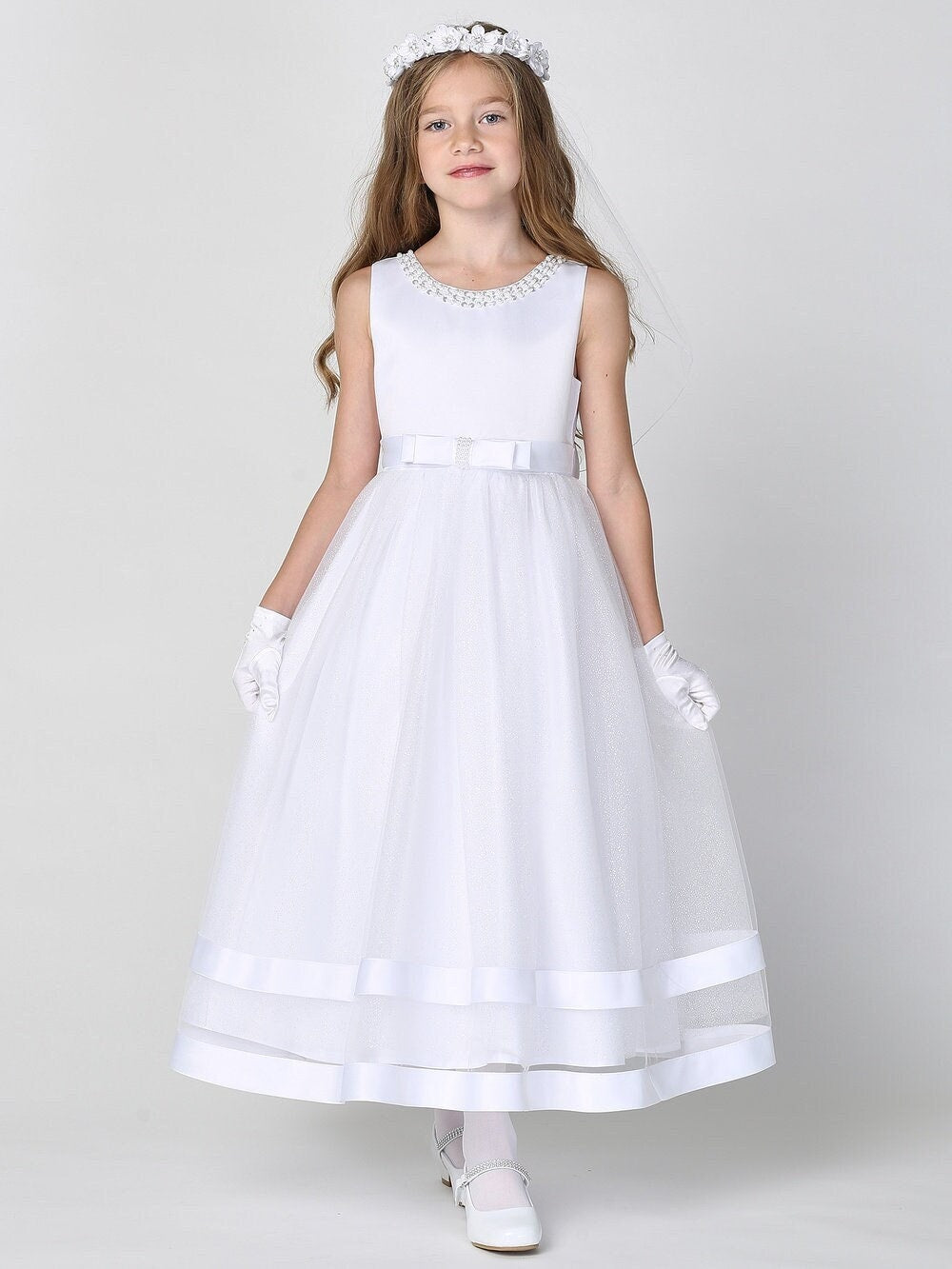 A girl wearing a graceful white First Communion Dress with a satin bodice and a glitter tulle skirt, adorned with a satin bow at the waist.