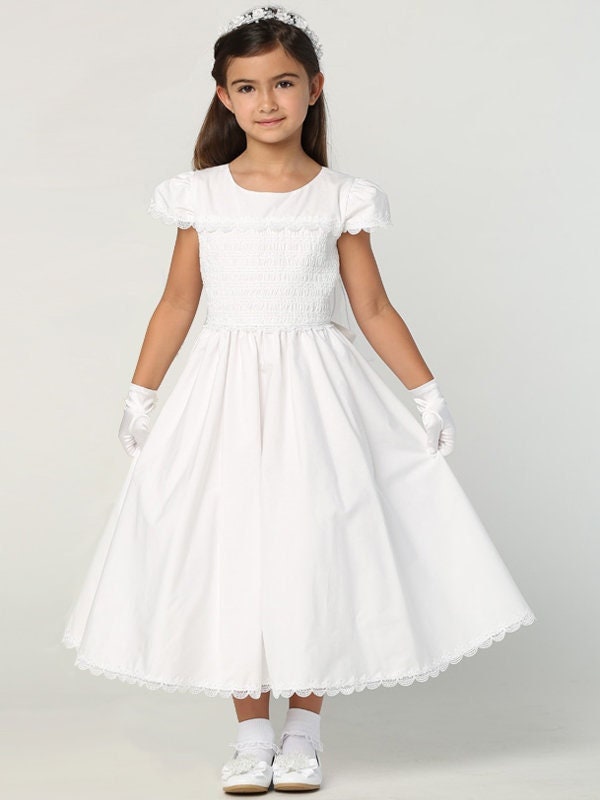 A girl wearing an elegant white First Communion Dress with a smocked cotton bodice and cotton skirt.