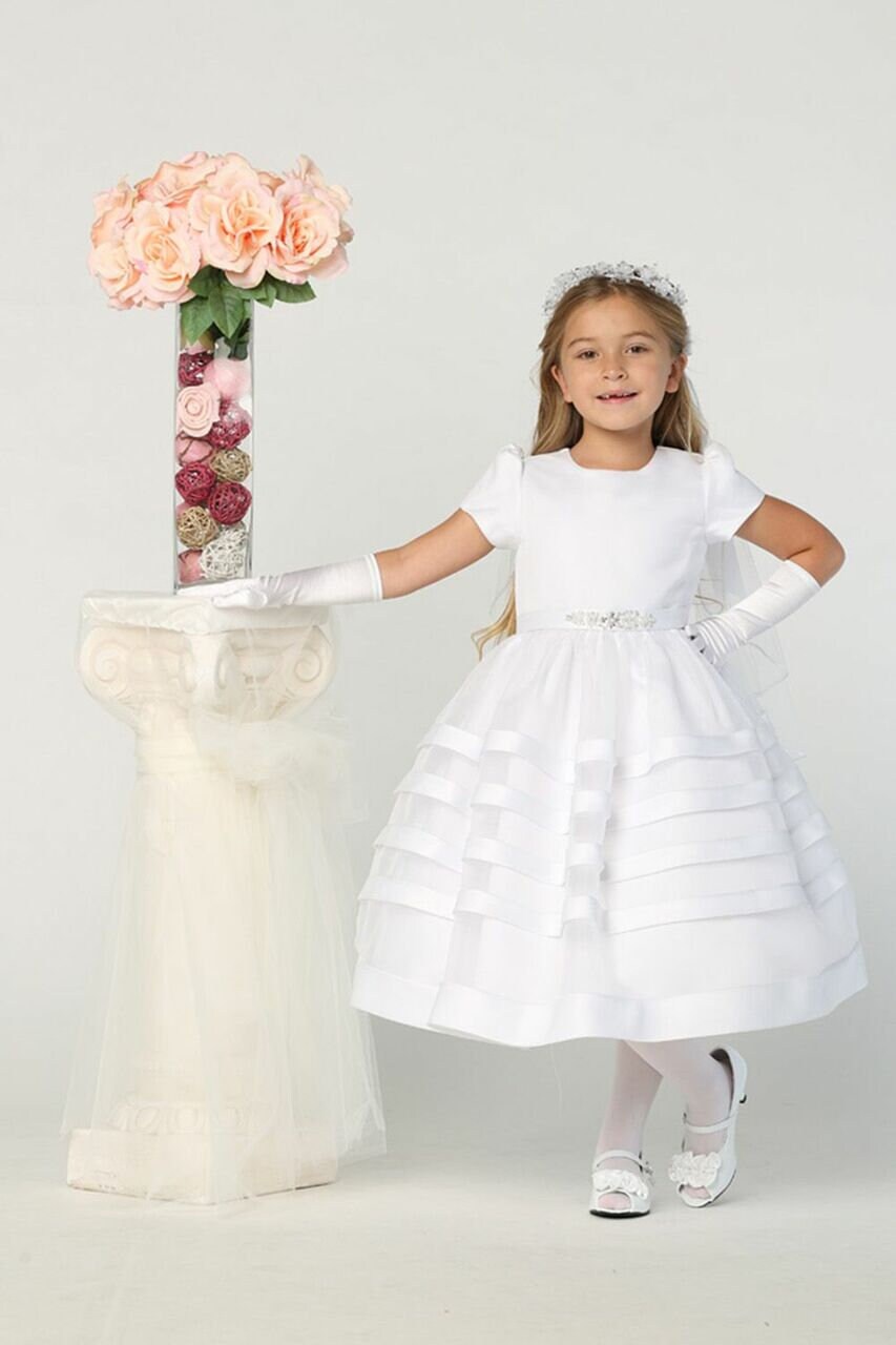 Girl wearing Elegant First Communion Dress with satin bodice and organza skirt, styled with gloves, floral headpiece, and white shoes.