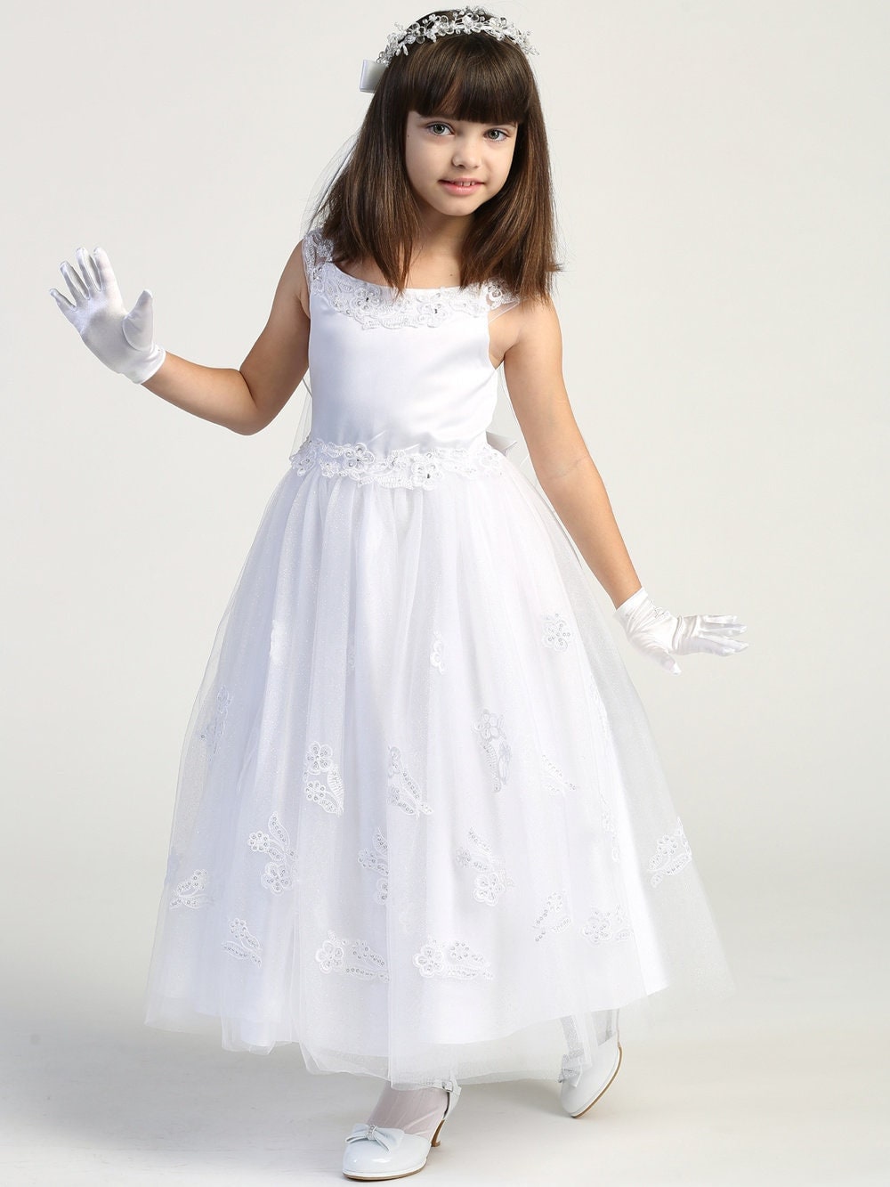 A girl wearing an elegant white First Communion Dress with a satin bodice adorned with embroidered appliques trimmed with beads and sequins.