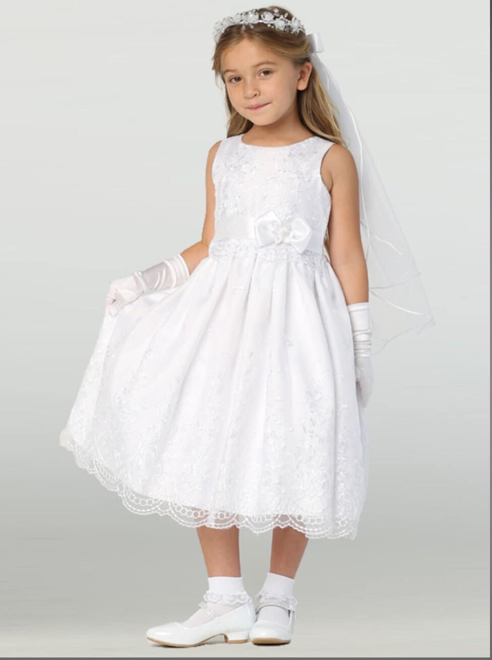 A girl wearing a beautiful white First Communion Dress with embroidered organza and satin ribbon trim.