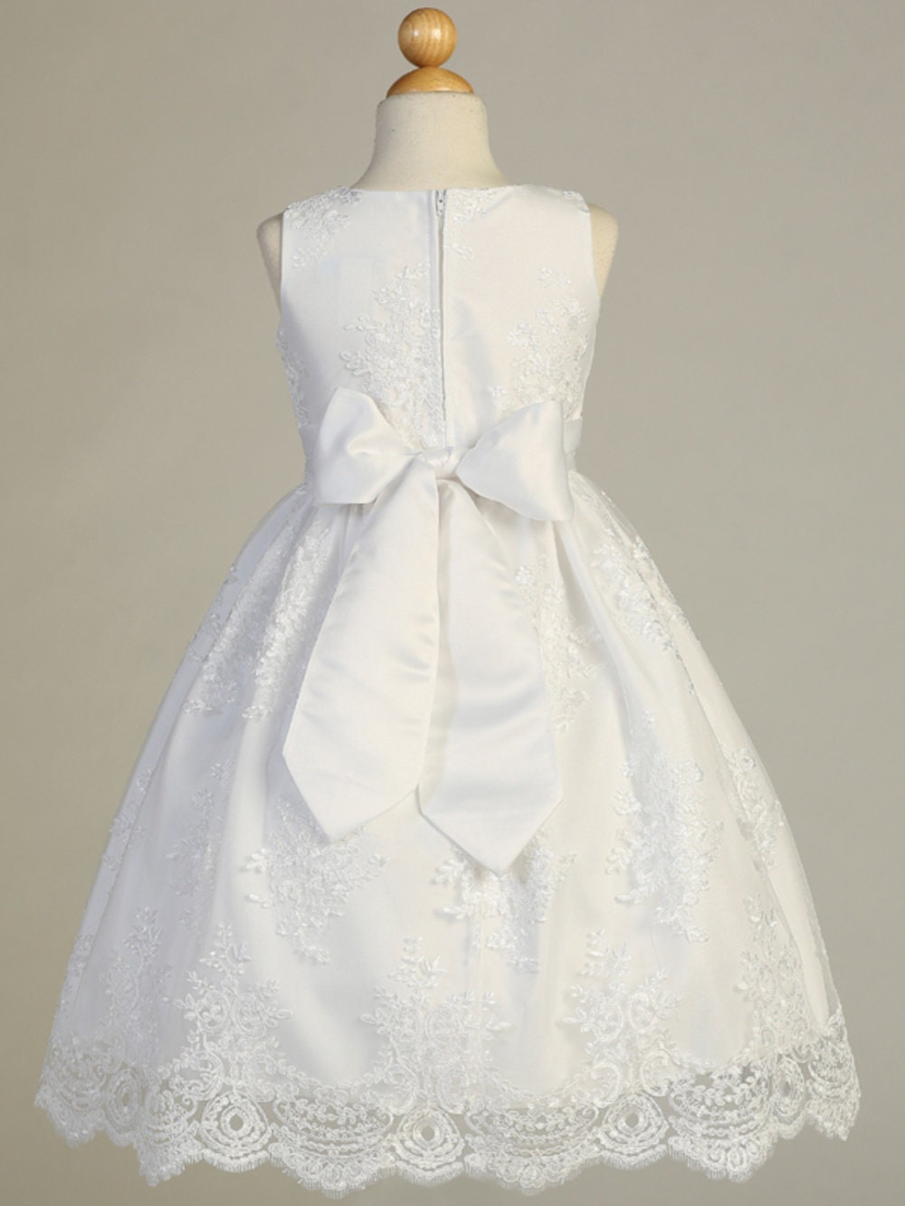 Back view of white First Communion dress featuring a large satin bow and intricate corded lace embroidery on a tea-length skirt.