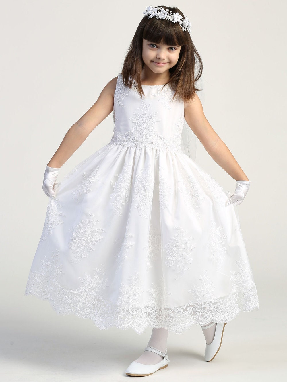Elegant white First Communion dress with corded embroidery lace, sleeveless bodice, and tea-length skirt, modeled by a young girl.